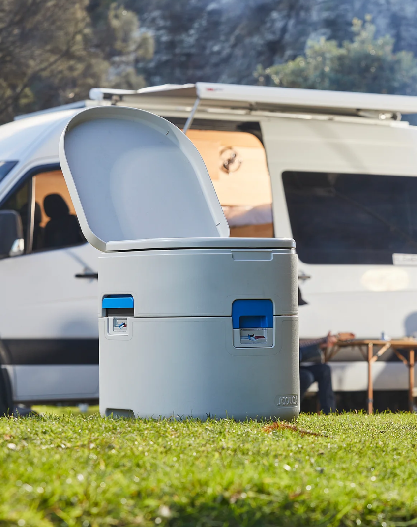 Portable outdoor composting toilet with open lid set on grass in front of a white camper van in a natural, wooded setting.