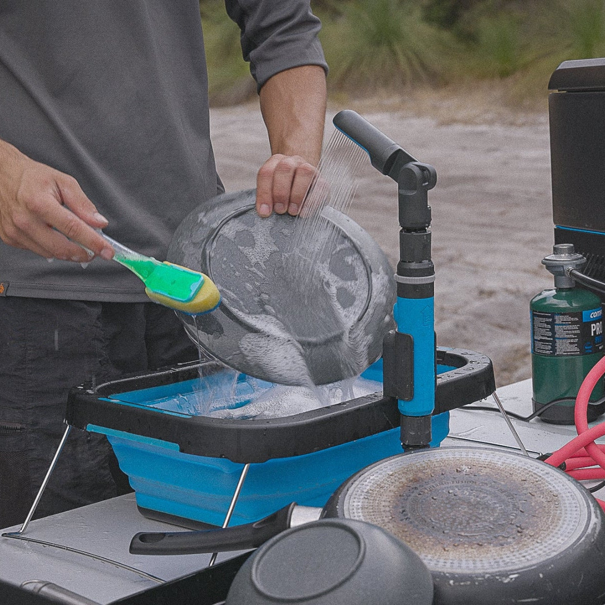 Camper washing a pan with a brush over the blue Joolca Flexi Sink outdoors, rinsing under a HOTTAP water heater spray