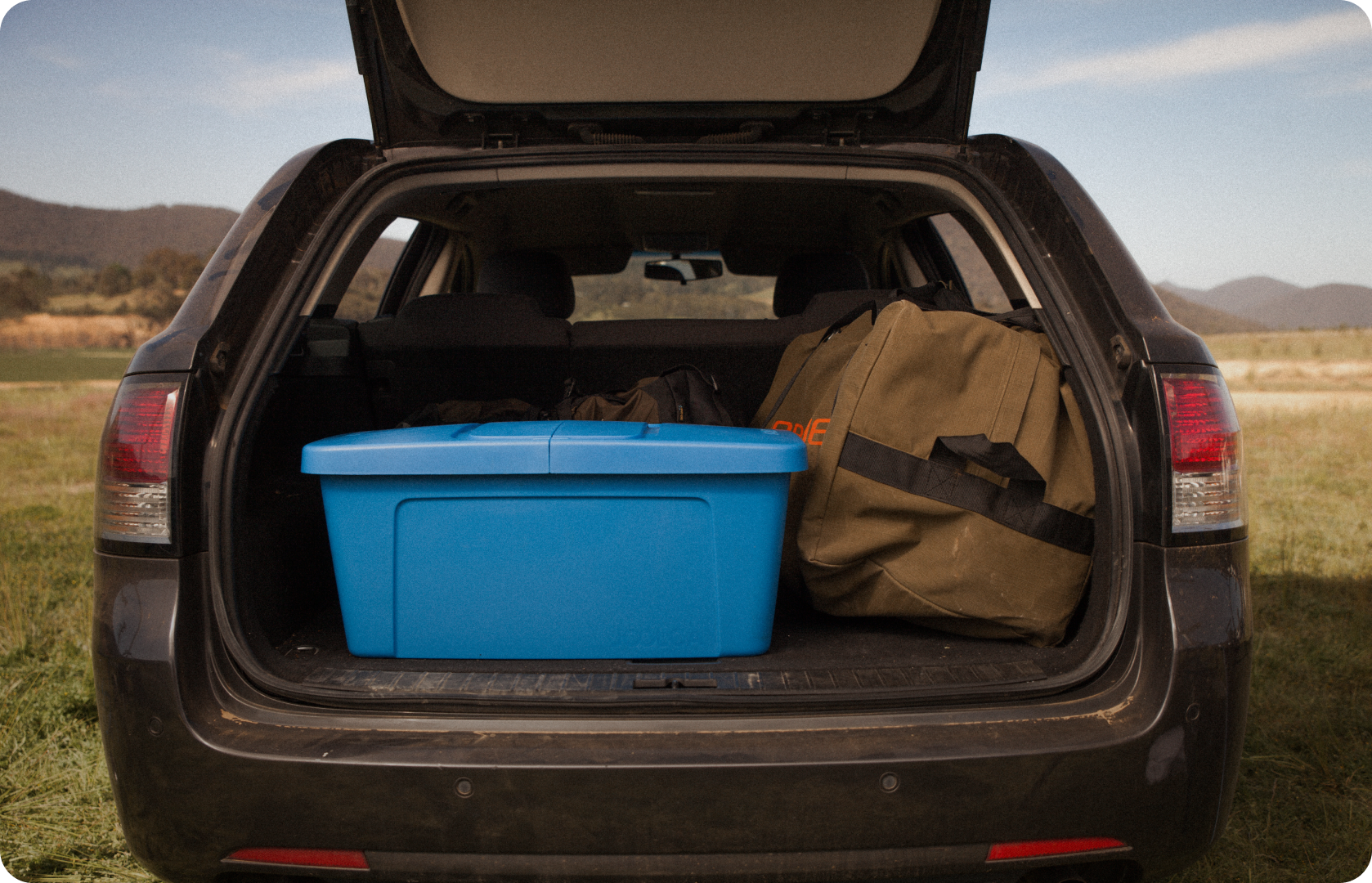 Open rear of a vehicle parked in a natural outdoor setting, with a large blue plastic storage bin and a green duffel bag packed inside the cargo area.