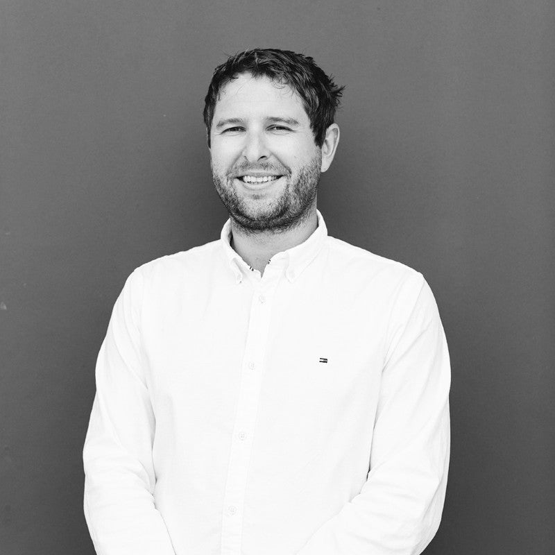 Smiling man with short hair and beard wearing a white button-up shirt standing against a solid dark background.