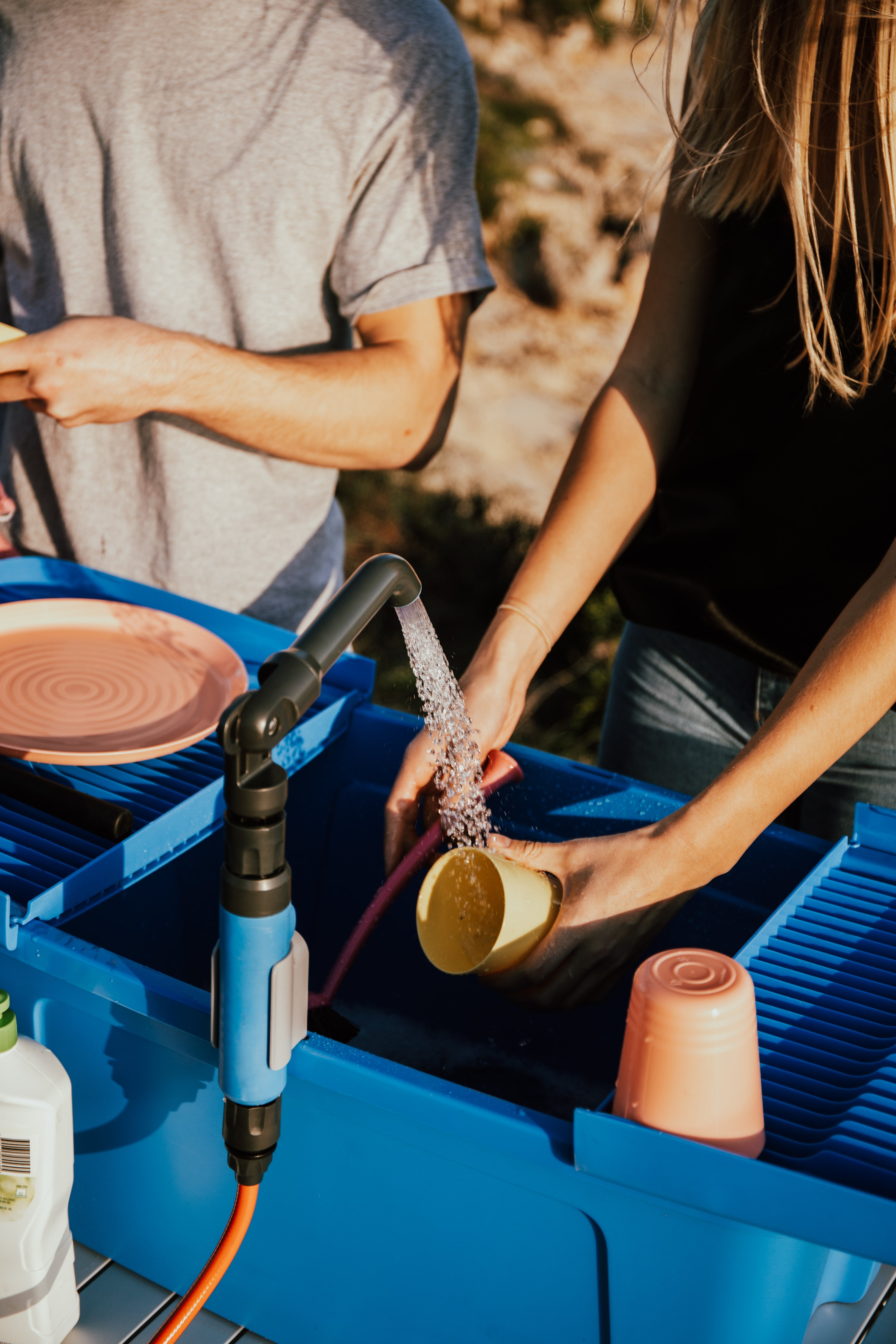 Two people using a portable outdoor water station with running water filling a cup, surrounded by dishwashing accessories and outdoor background.