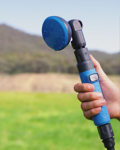 Hand holding the HOTTAP V2 showerhead outdoors, thumb resting on the on-off button in the handle.